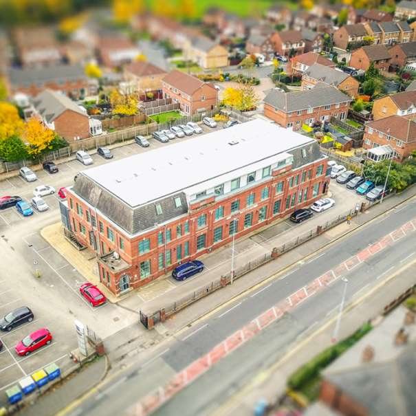Aerial view of a rectangular brick office building with a white roof, adjacent parking lot with cars, and surrounded by residential houses and trees—one of the notable Morson Projects locations.