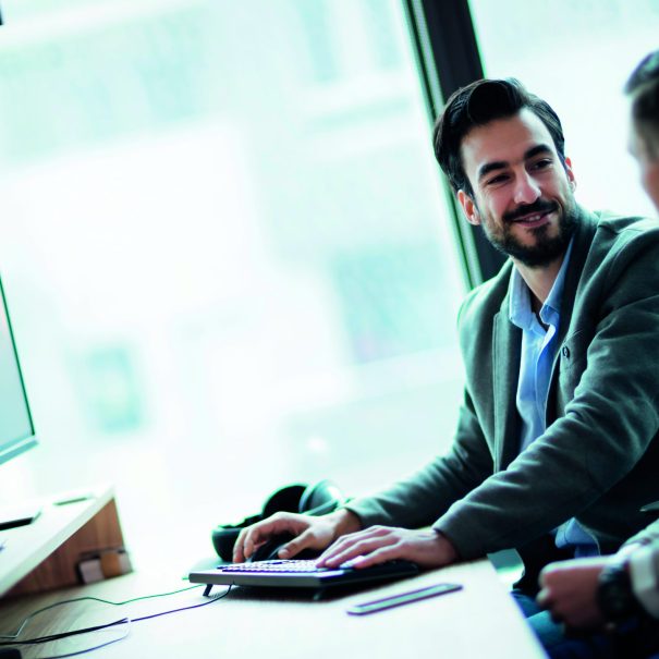 Two men in business attire sit at a desk with a computer, engaged in conversation about data translation in a modern office setting.