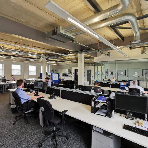 Modern open-plan office with several people working at desks, computers, and monitors; exposed ceiling ducts and large windows provide natural light.