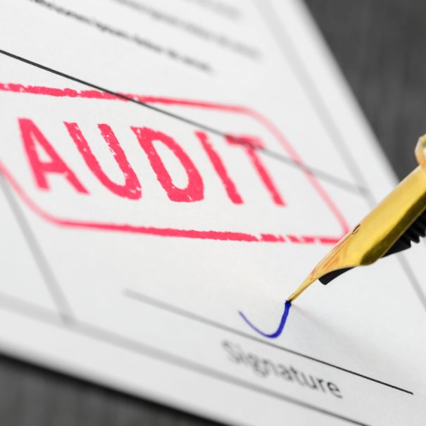 A close-up of a document stamped "AUDIT" in red, with a person signing the signature line using a fountain pen.