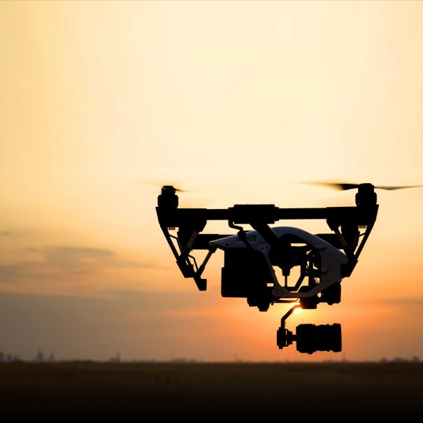 A drone with a mounted camera flies at sunset, silhouetted against the sky over an open field.