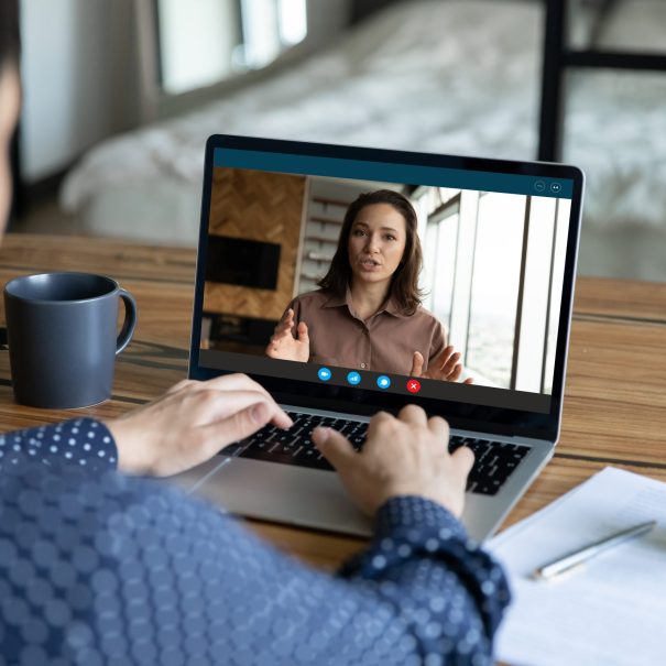 Person participating in a video call on a laptop, with a woman speaking on the screen; a mug, notebook, and pen are on the desk.