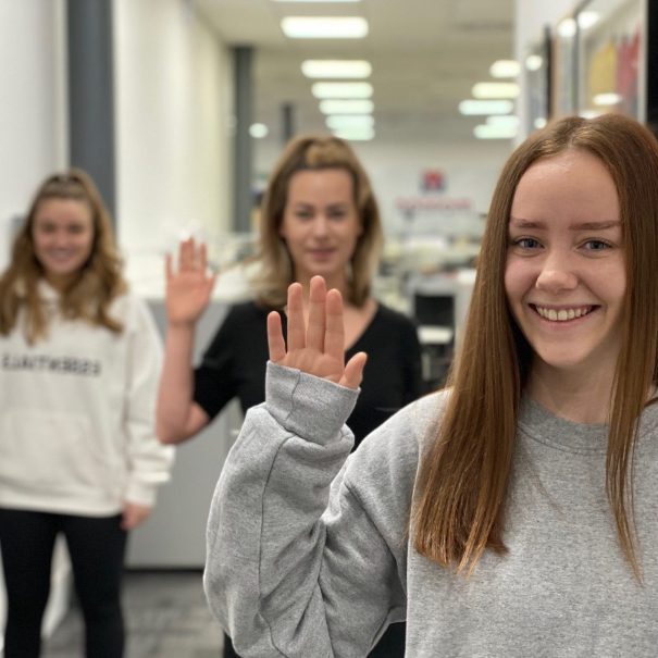 Three women standing in a hallway indoors, each holding up their right hand in a wave, with the focus on the woman closest to the camera.