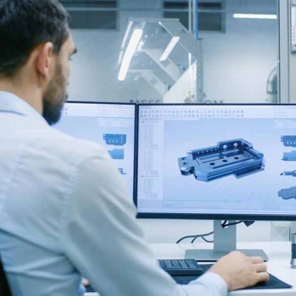 A man sits at a desk working on a computer displaying a 3D modeling software with a technical component design.