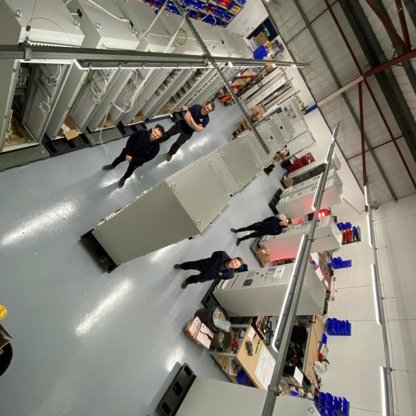 Industrial workshop with several large metal cabinets, shelving with parts along the walls, and six people standing at intervals in the central aisle.