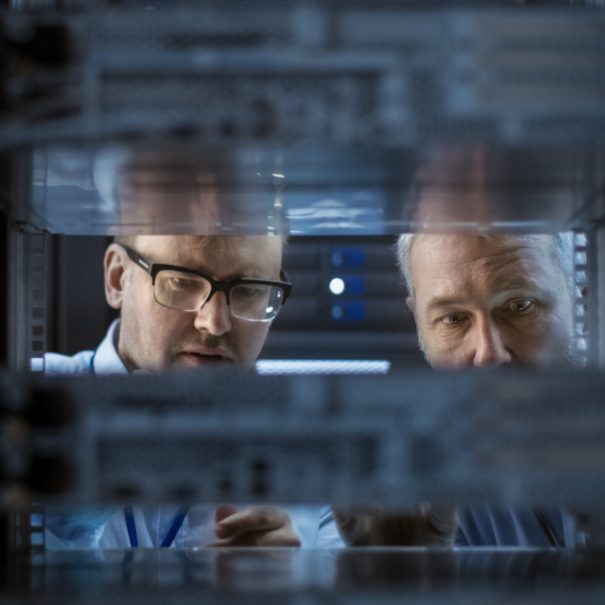 Two men examine server hardware inside a data center rack, viewed through the metal frame and electronic components.