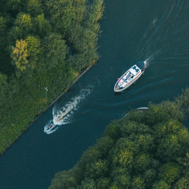 Aerial view of a narrow river bordered by dense trees, with a small motorboat and a larger yacht traveling in opposite directions.