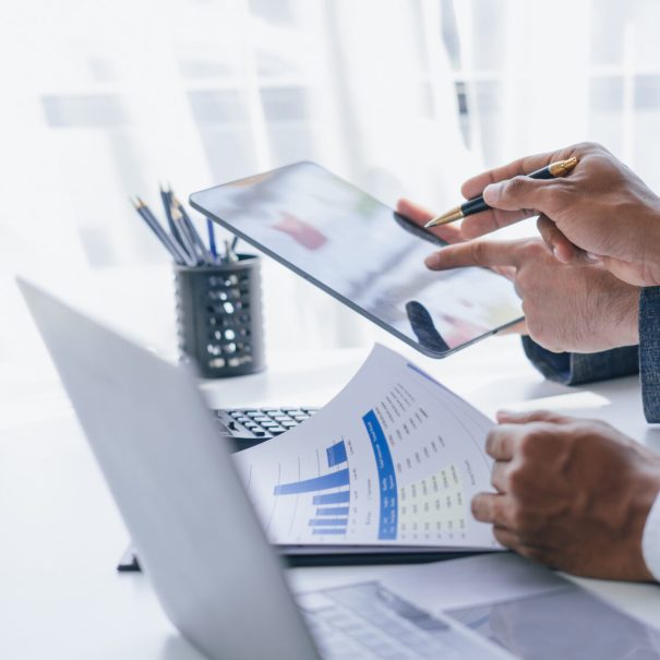 Two people in business attire review a financial chart on paper and a tablet, seated at a desk with office supplies and a laptop displaying CNC programming data.