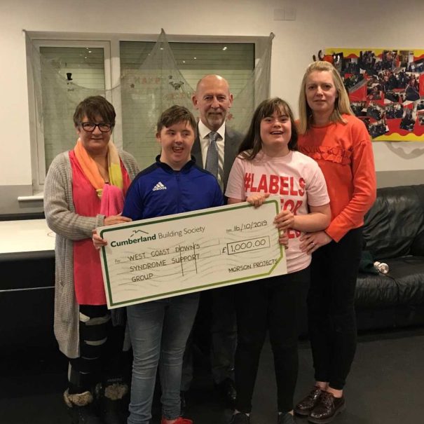 Five people stand indoors holding a large check for £1,000 made out to "West Coast Down's Syndrome Support Group" from Cumberland Building Society.