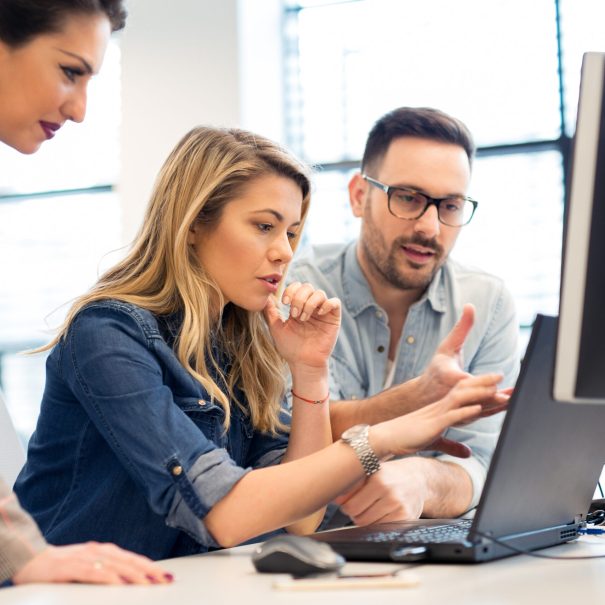 Three people work together at a desk, looking at a computer monitor and discussing something on the screen in an office setting.