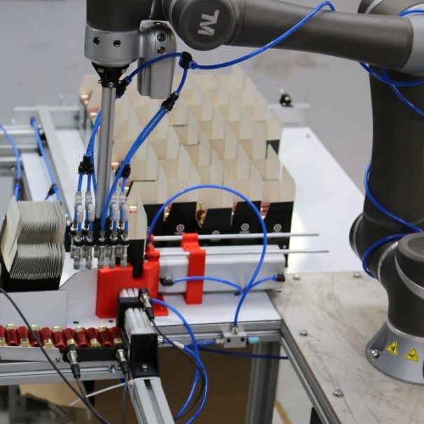A robotic arm equipped with blue pneumatic tubes operates on an automated assembly line with various mechanical and electronic components.