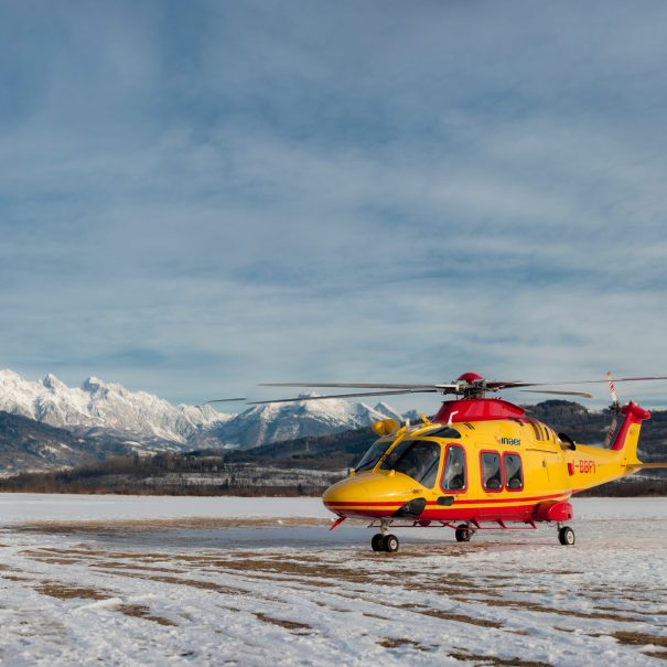 A yellow and red rescue helicopter is parked on a snowy field with mountains visible in the background under a partly cloudy sky.