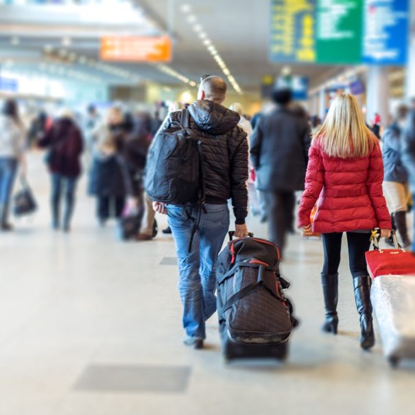 A man and a woman walk through a busy airport terminal, each pulling wheeled suitcases, with other travelers visible in the background.