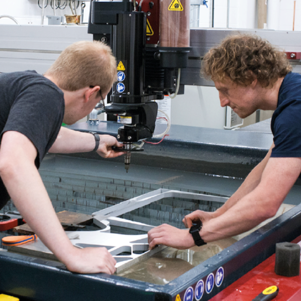 Two people operate a CNC machine, closely inspecting and adjusting a metal sheet being cut inside an industrial workshop.