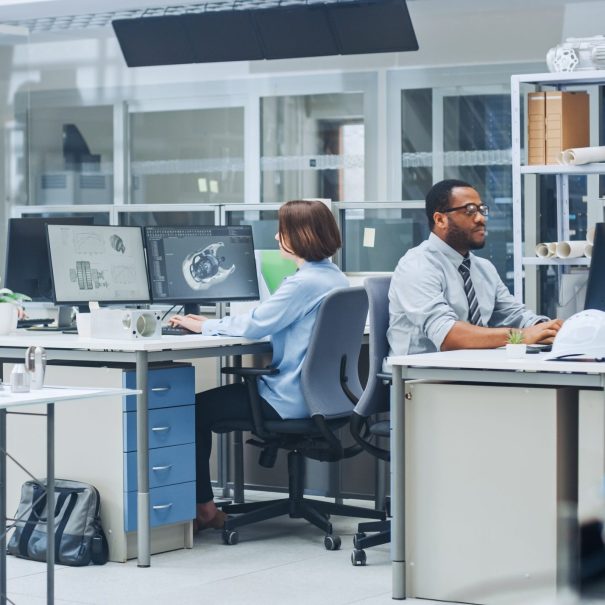 Three people work at desks with computers in a modern office, focusing on engineering or design tasks; various equipment and models are visible on the desks and shelves.