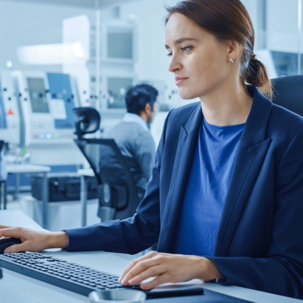 A woman in business attire works at a desktop computer in a modern office with other people in the background.