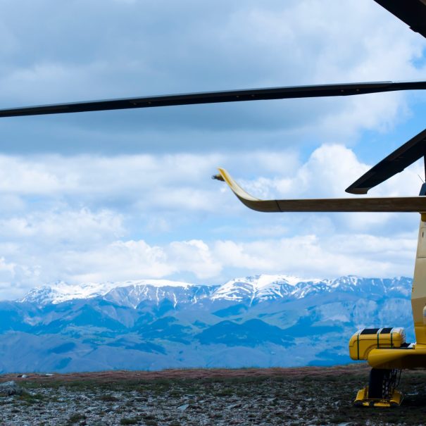 A yellow helicopter is parked on rocky ground with snow-capped mountains and a cloudy sky in the background.