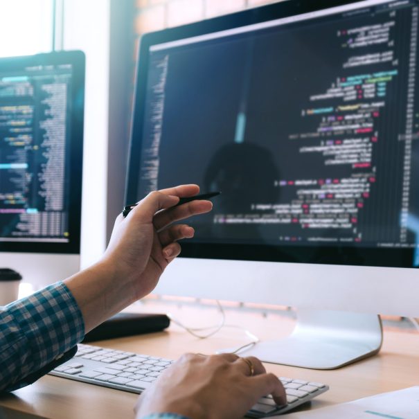 A person points at lines of code on a computer monitor while working at a desk with two screens and a keyboard.