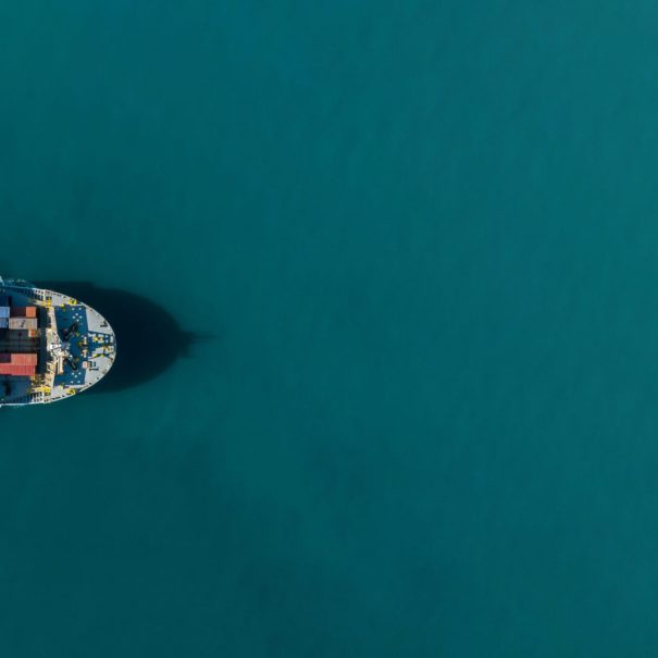 A cargo ship carrying colorful containers moves through calm blue water, viewed from above.