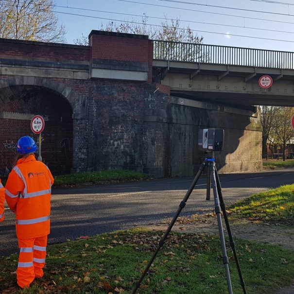 Two workers in orange safety gear and blue helmets stand near a tripod-mounted device, facing a brick railway bridge over a road with clearance signs.