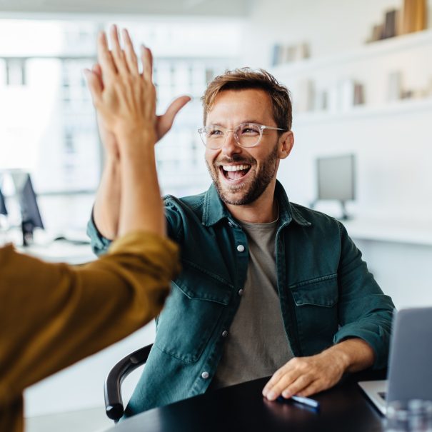 Two people sitting at a desk in an office share a high five while a third person smiles nearby; a laptop and office supplies are on the table.
